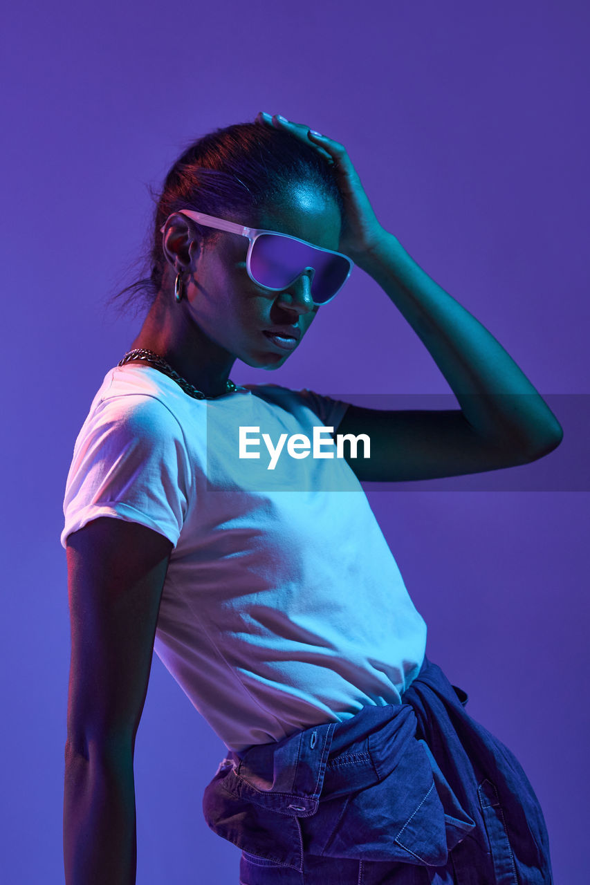 Stylish brazilian female in fashionable sunglasses and outfit touching hair while standing on purple background in studio with fluorescent light