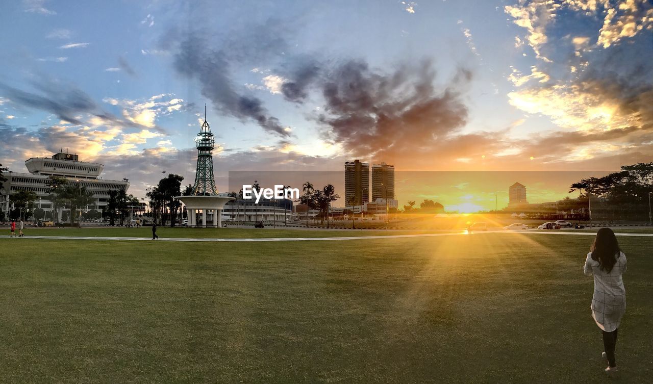 Rear view of woman walking on grass in city against sky during sunset