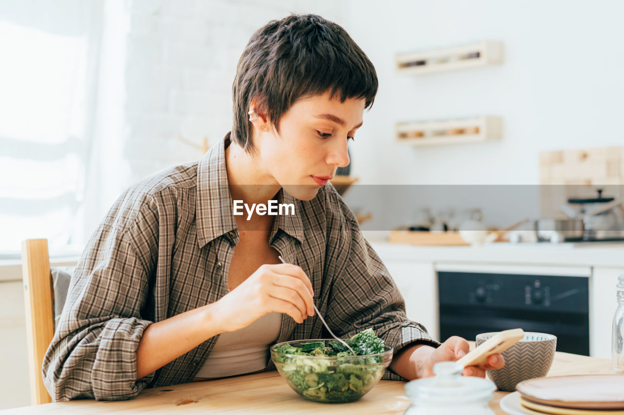 A young short-haired brunette  in the kitchen eating and browsing social networks on her phone.