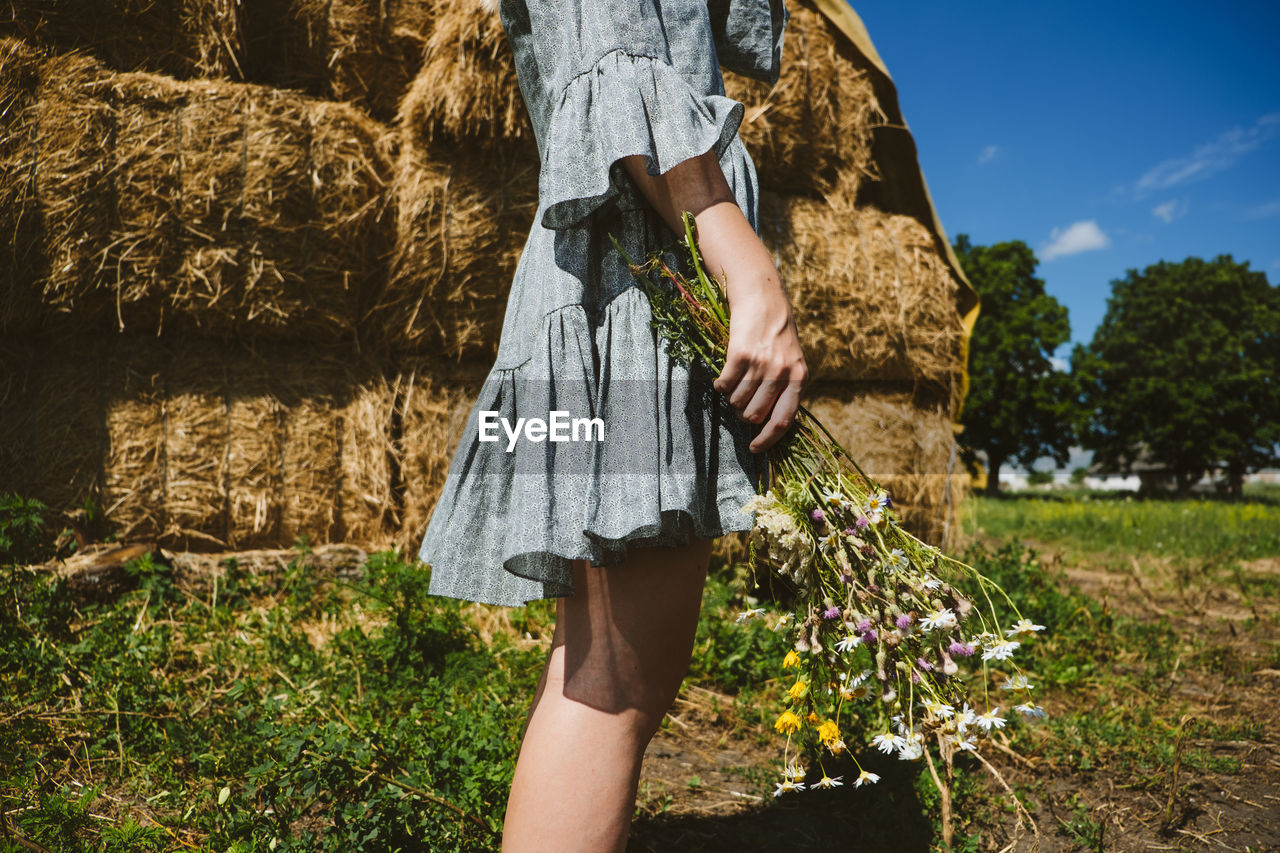 Young girl in rubber boots with flowers standing against the background of straw bales on country 