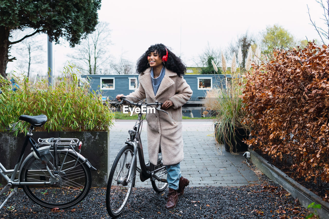 Smiling young african american female with curly dark hair wearing warm coat walking with bicycle while listening to music in headphones on street in autumn day