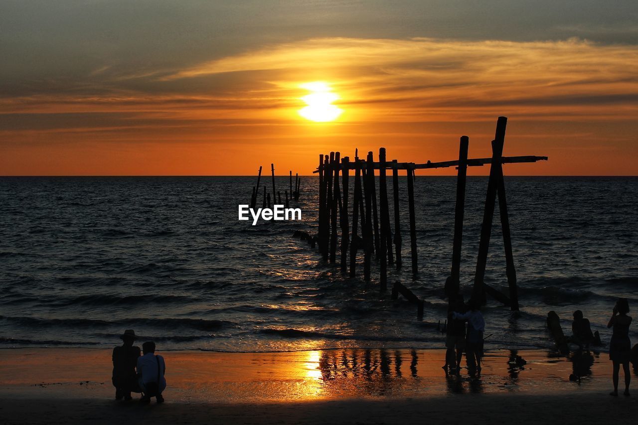 SILHOUETTE PEOPLE ON WOODEN POST ON BEACH AGAINST SKY DURING SUNSET