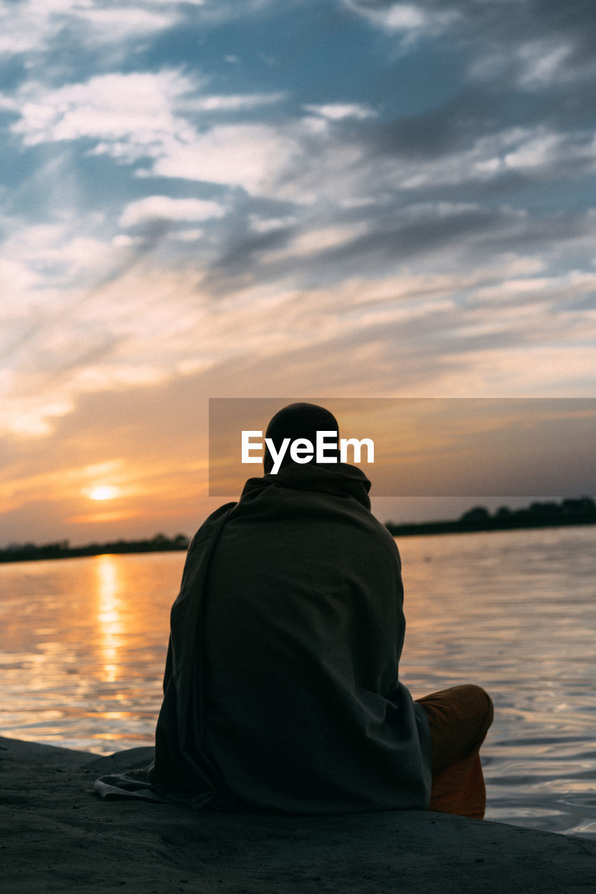 REAR VIEW OF MAN SITTING ON SEA AGAINST SKY DURING SUNSET