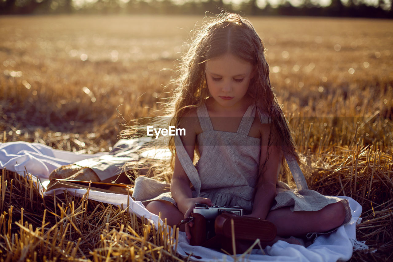 Girl child in a dress sitting on a mown field with a camera on a blanket with bread and a book