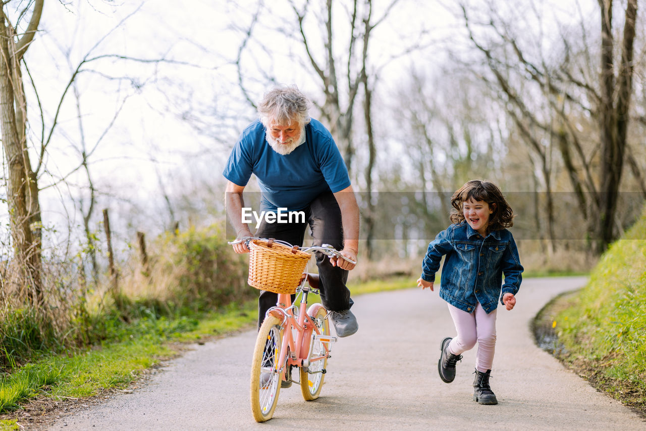 Full body of active grandfather riding bicycle of granddaughter near positive running girl on path in countryside with green trees