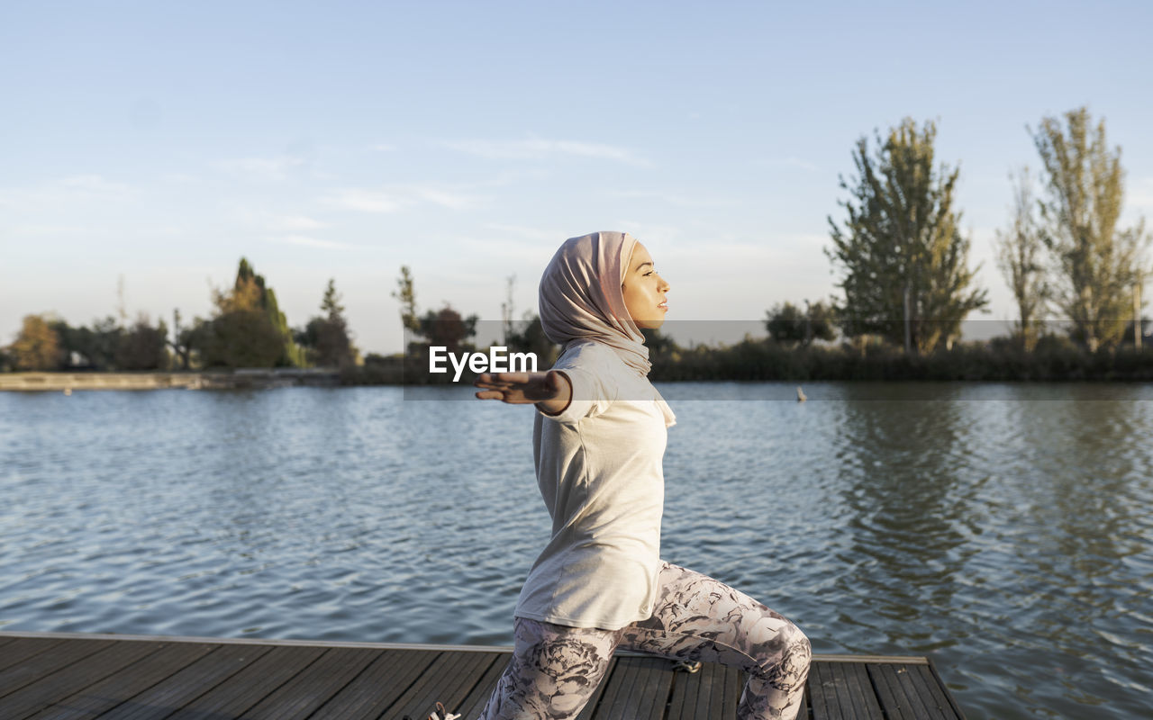 Side view of arab female in hijab doing crescent lunge twist on knee asana with spread arms on waterfront near rippling river