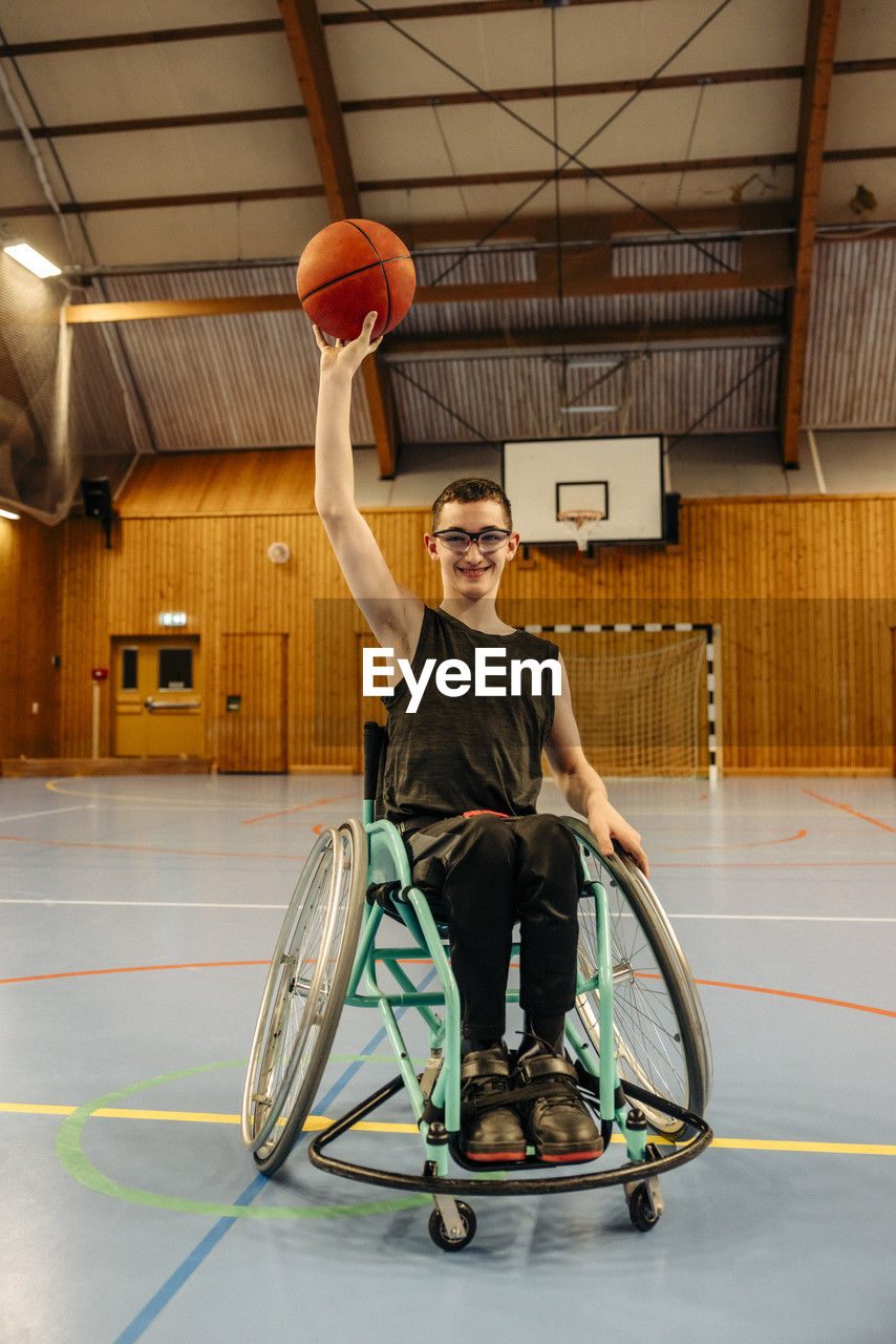Smiling girl balancing basketball while sitting on wheelchair at sports court