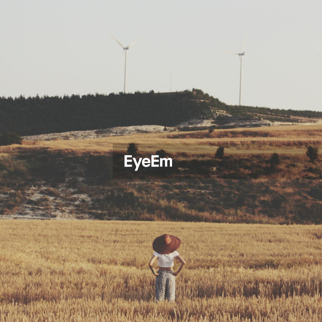 Woman standing in field of wheat and looking at hill with wind turbines
