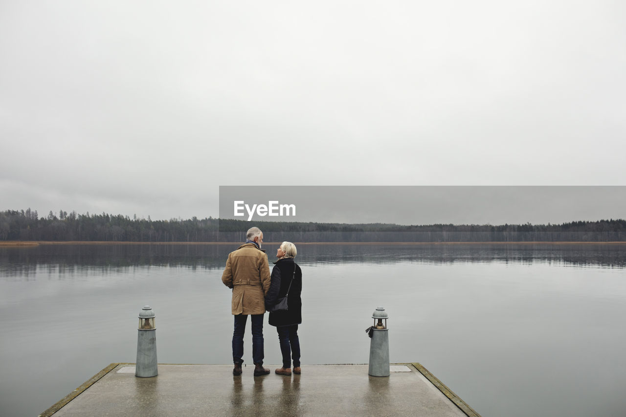 Rear view of senior couple talking while standing by lake against clear sky