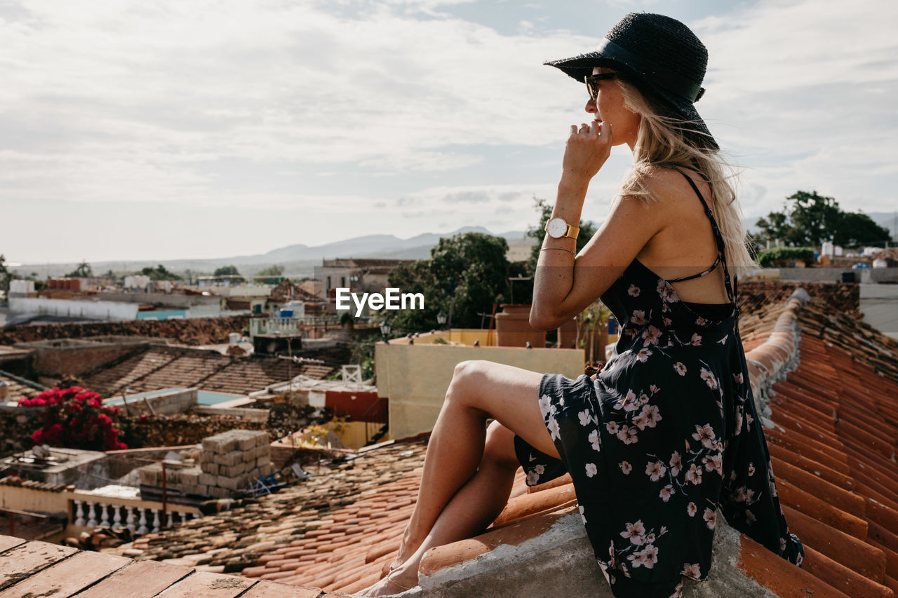 Side view of young woman looking at cityscape while sitting on rooftop against sky during sunny day
