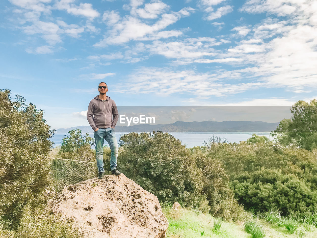 WOMAN STANDING ON ROCK AMIDST PLANTS AGAINST SKY