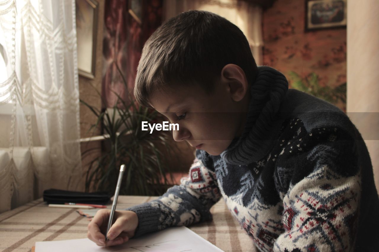 A boy in a christmas sweater sits at the table and draws and writes