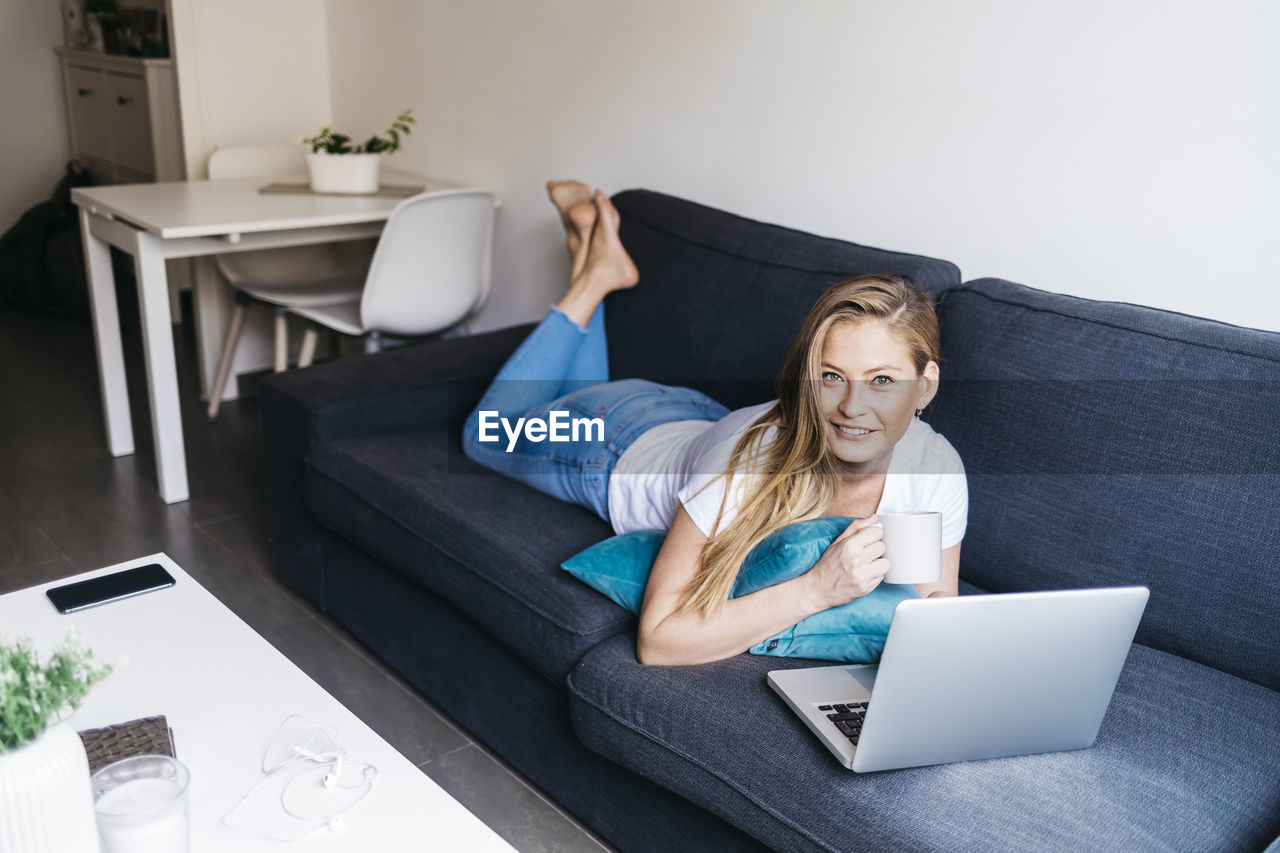 Portrait of young woman using laptop while lying on sofa at home