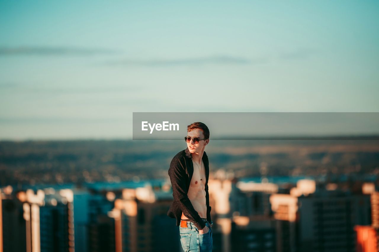 PORTRAIT OF YOUNG MAN STANDING AGAINST CITYSCAPE AGAINST SKY