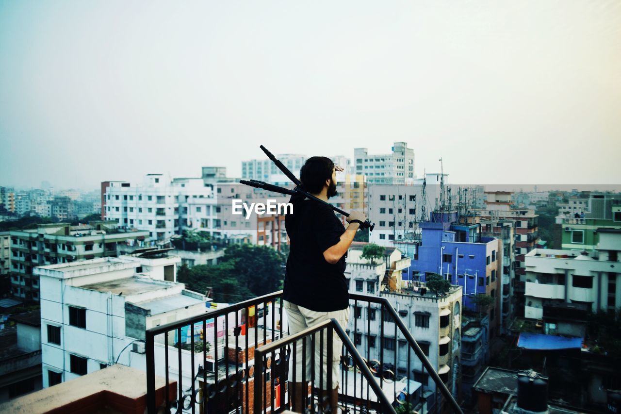 Side view of man holding tripod while standing on terrace in city