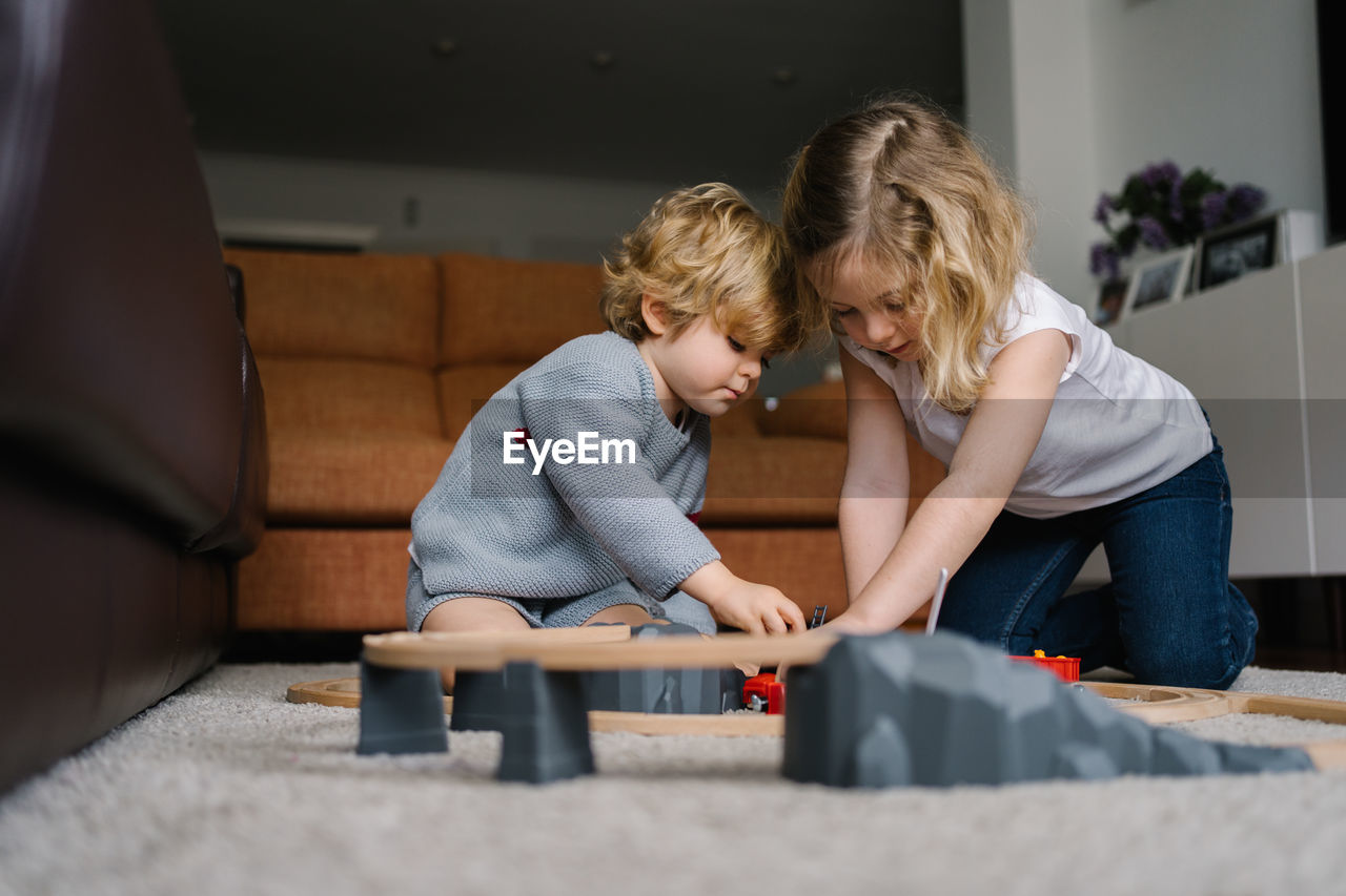 Cute little curly haired siblings sitting on floor and playing with toy road and cars while spending time together in living room at home