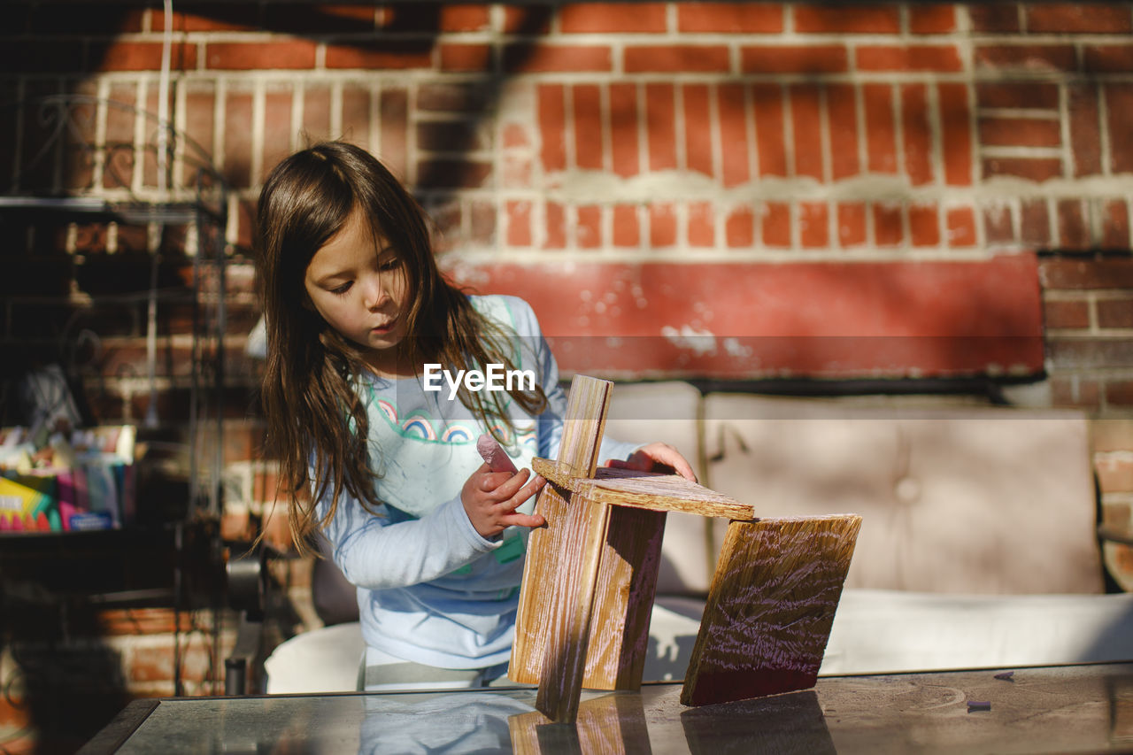 A focused child concentrates on building with wood against brick wall