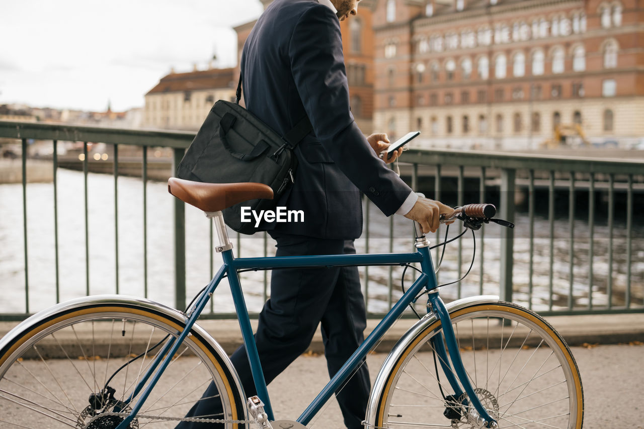 Midsection of businessman using smart phone while walking with bicycle on bridge in city