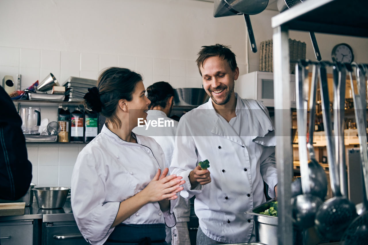 Male chef holding broccoli while talking with female colleague at kitchen