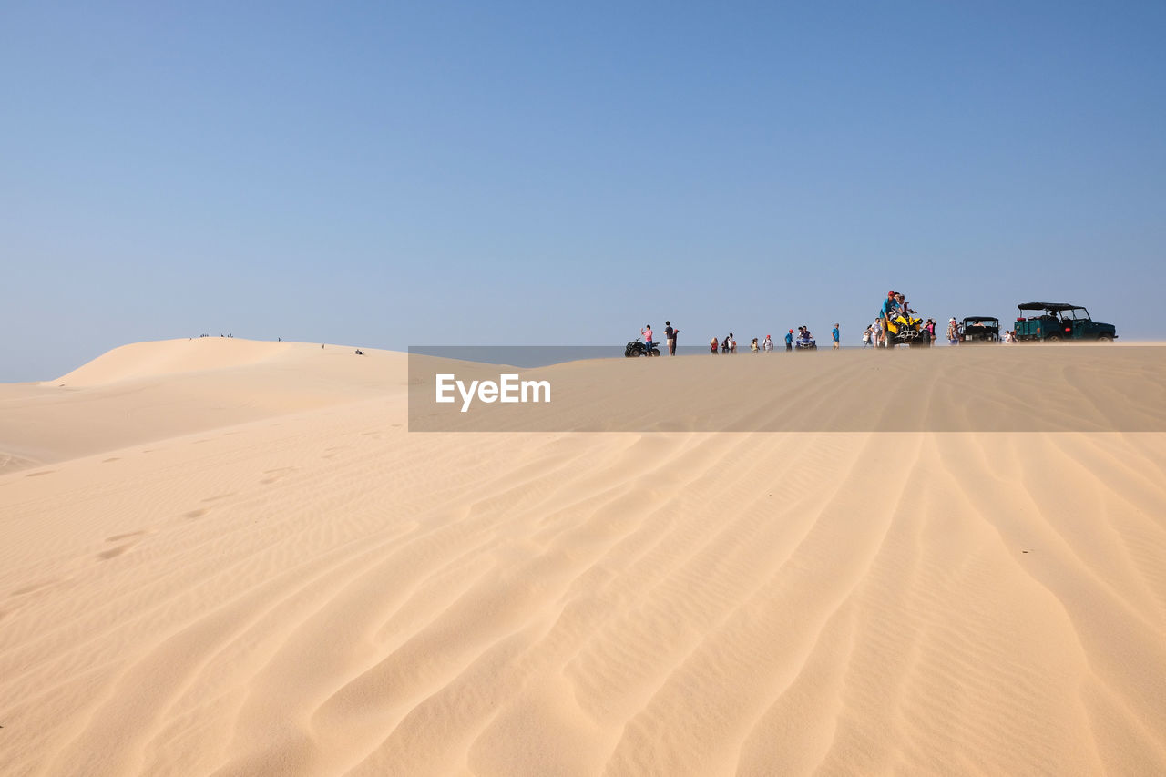 Low angle view of people with vehicles on sand dune in desert against blue sky