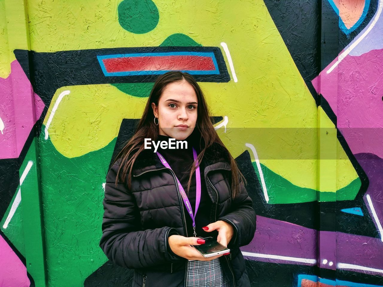 Portrait of young woman holding mobile phone against colorful graffiti wall