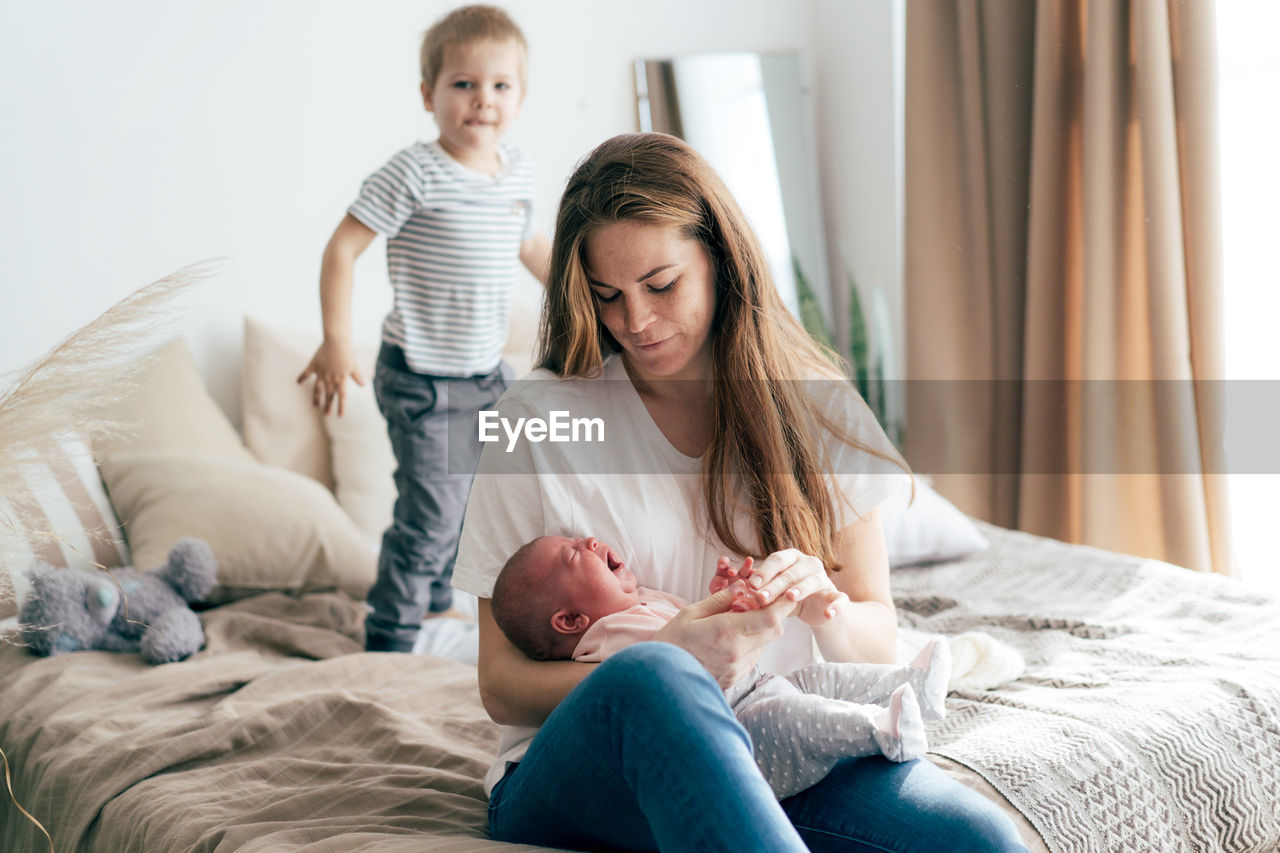 Young caucasian mother with newborn baby and toddler son on bed at home.