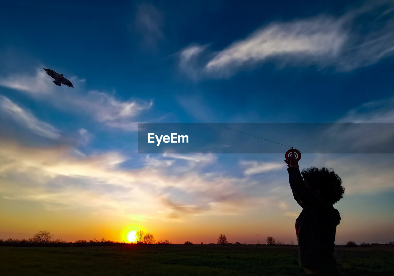 Silhouette kid flying kite in sky during sunset