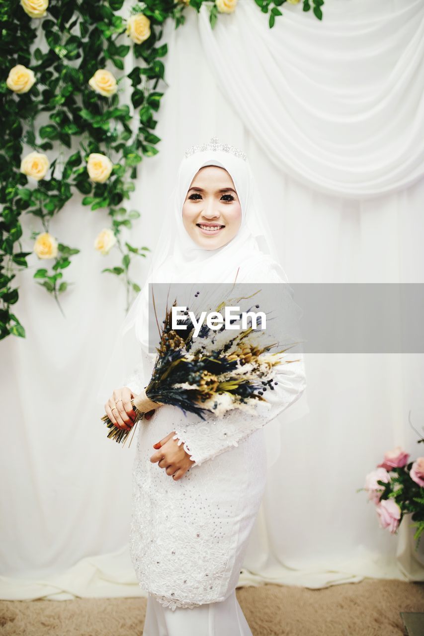 Portrait of young woman wearing hijab holding flowers while standing against curtain