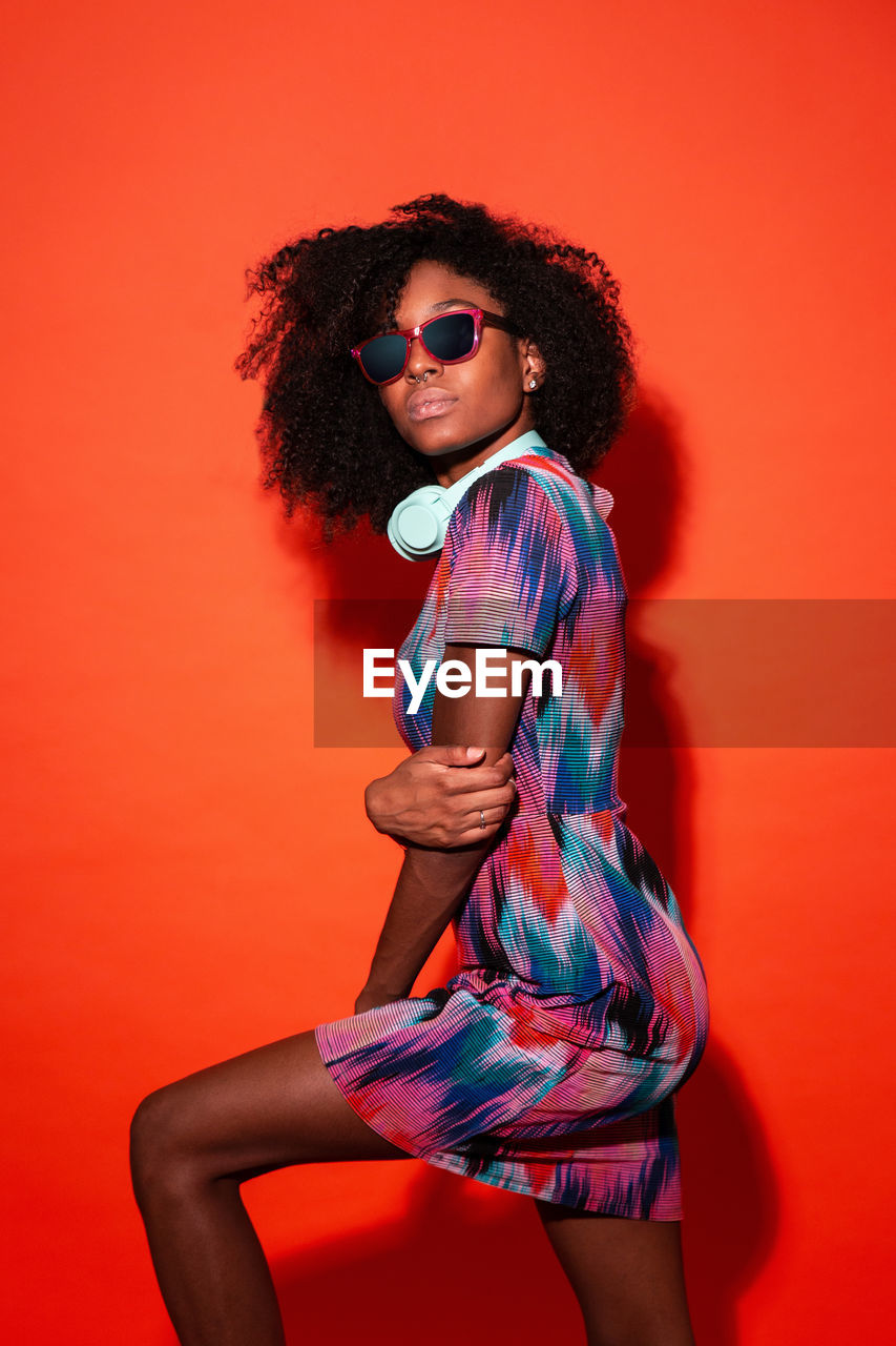 Modern cuban woman with afro hair wearing bright dress and sunglasses and headphones in red studio background