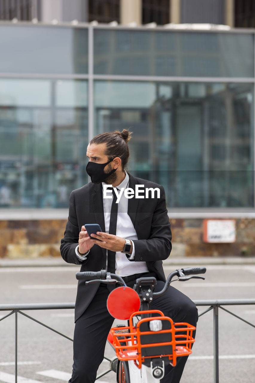 Portrait of businessman wearing protective face mask sitting on bicycle with smart phone in hands