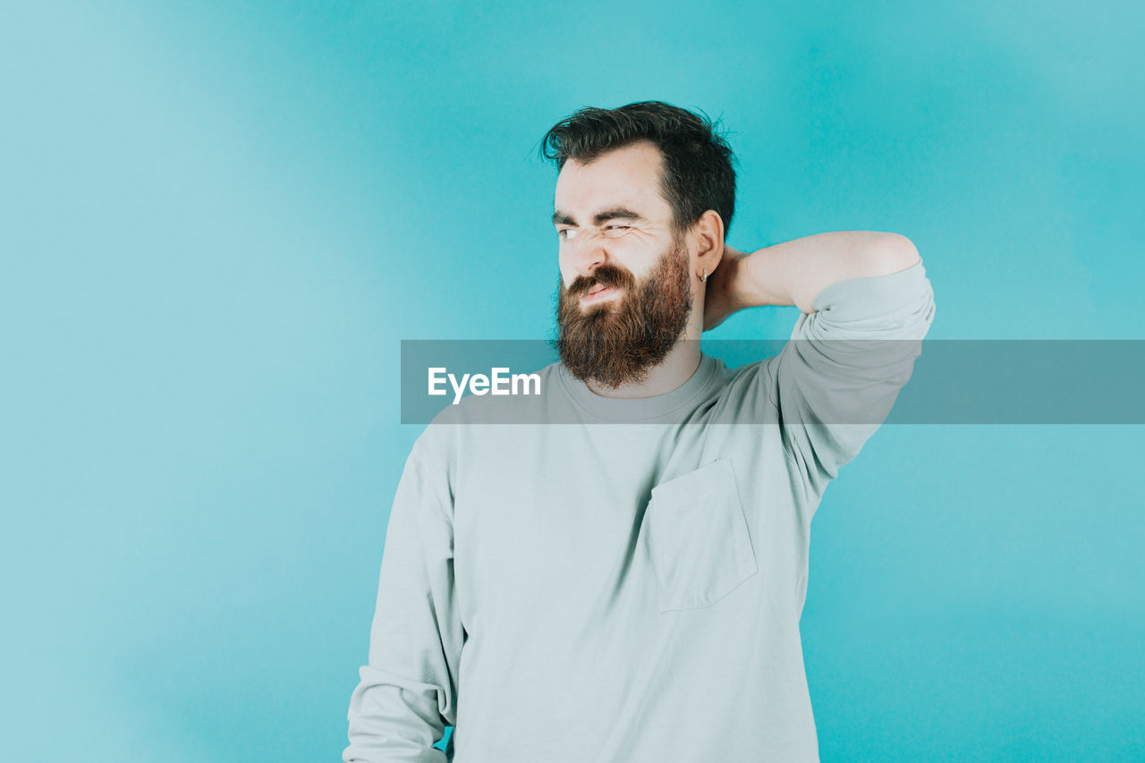 one person, beard, blue, facial hair, adult, men, colored background, studio shot, waist up, portrait, blue background, indoors, person, young adult, copy space, standing, casual clothing, photo shoot, front view, looking, clothing, human face, lifestyles, brown hair, emotion, stubble, white, contemplation, looking at camera, smiling, turquoise