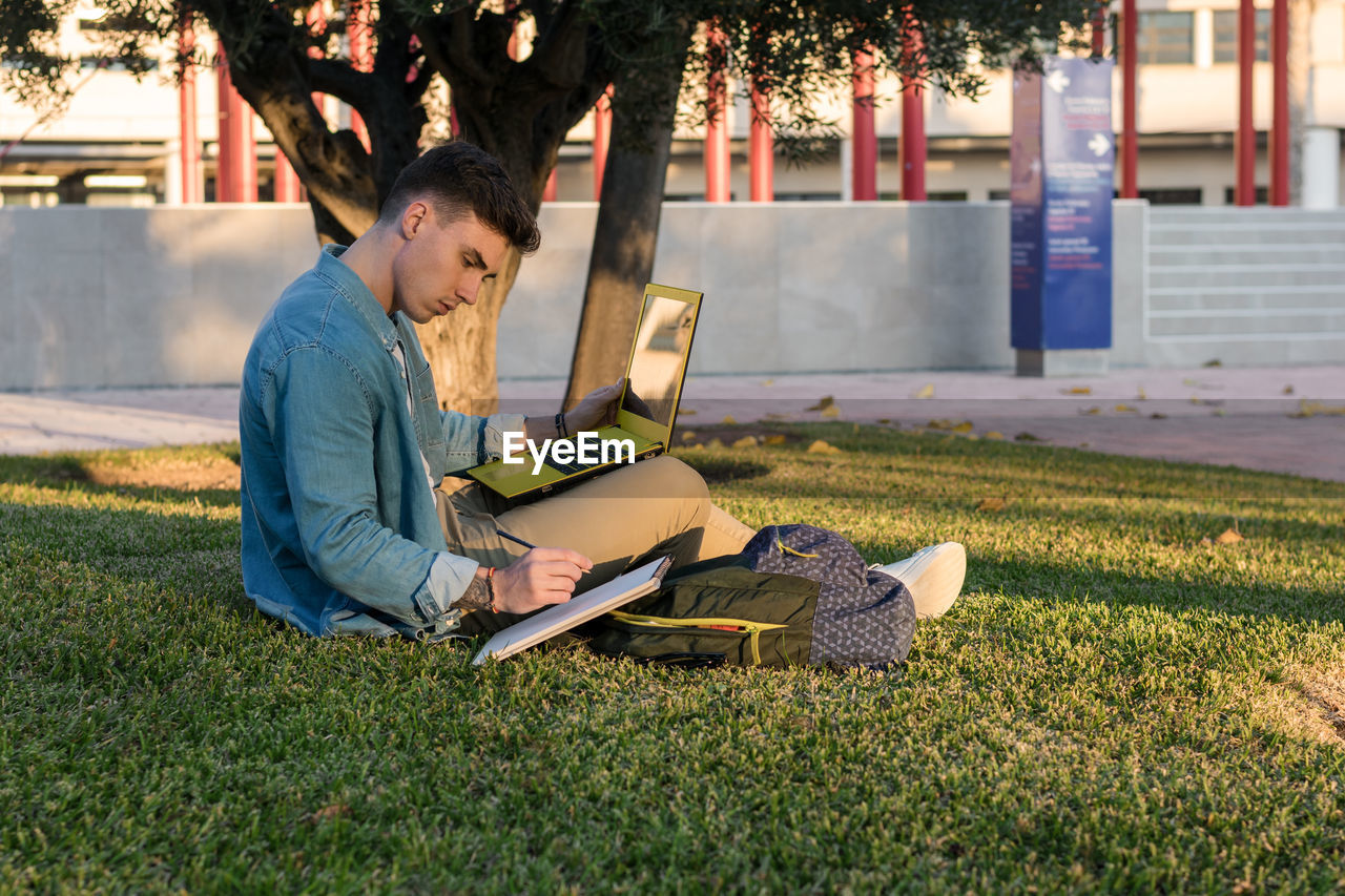 Smart concentrated man with backpack studying at laptop and writing on a notepad sitting in park grass with crossed legs in sunny day
