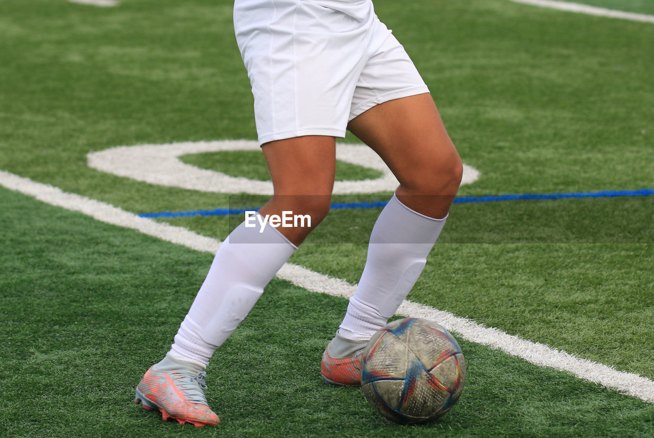 Close up of legs of a young soccer player in a white uniform with the ball and a green turf field.