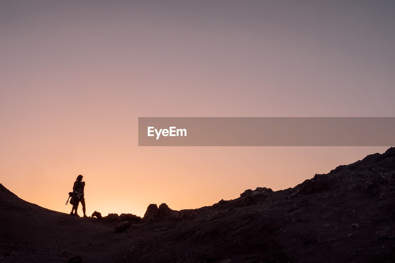 SILHOUETTE MAN STANDING ON ROCK AGAINST SKY AT SUNSET