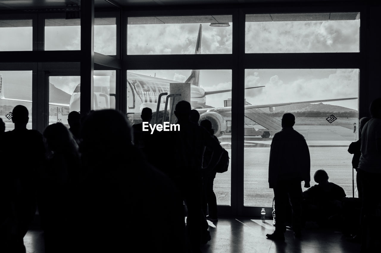 SILHOUETTE PEOPLE STANDING BY WINDOW AT AIRPORT