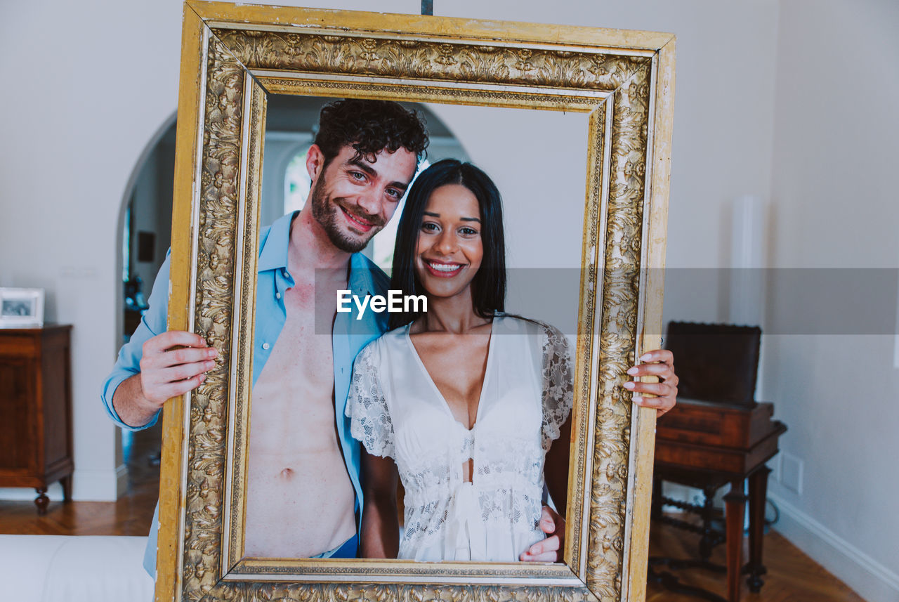 Portrait of smiling young couple holding picture frame at home