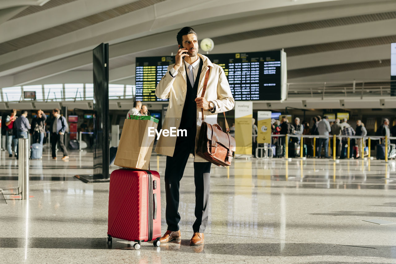 Man with dark hair in stylish clothes talking on phone while standing with suitcase in terminal of airport