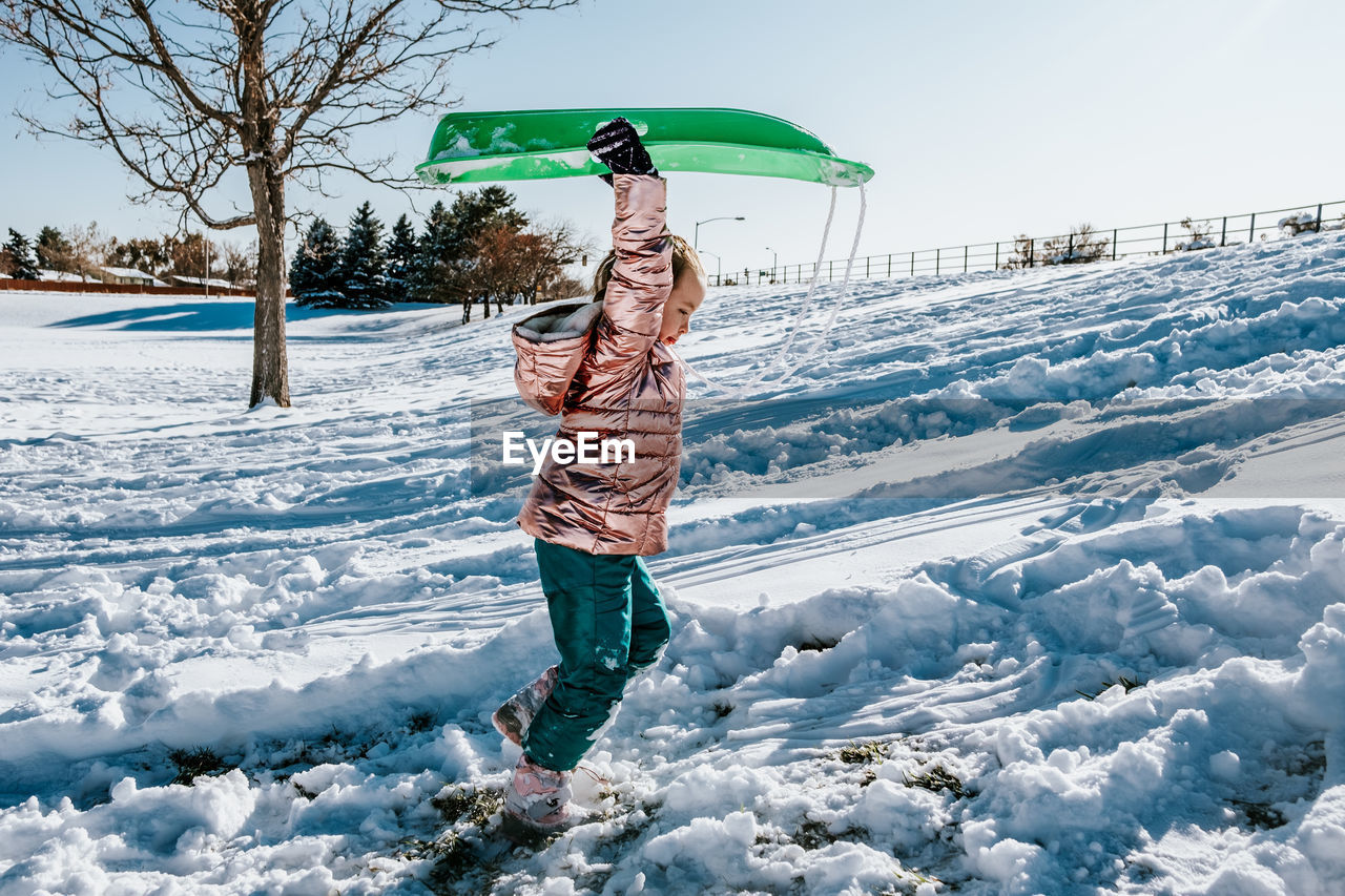 Side portrait of young girl carry sled up a snowy hill