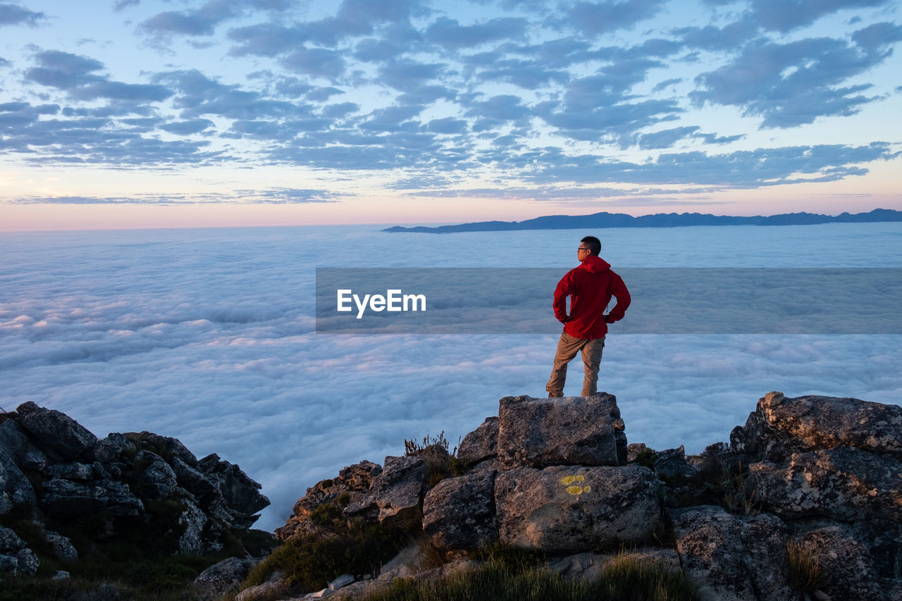 Man in red jacket  standing on mountain rock looking at clouds below 