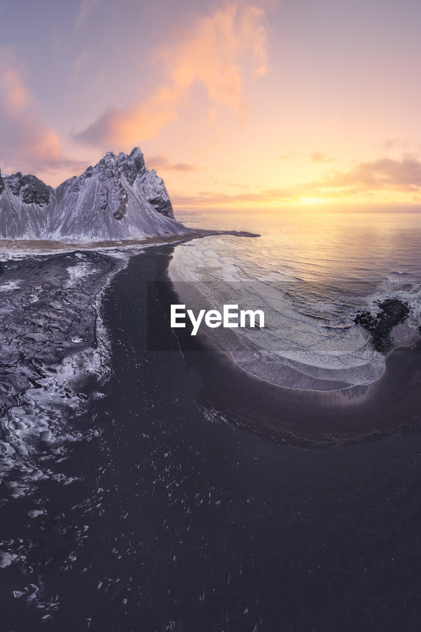 Drone view of calm frozen endless sea surrounding high rocky snowy vestrahorn mountain during picturesque sunset in stockness beach, iceland