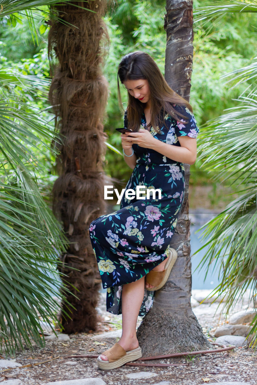 Full length of woman using mobile phone while standing by tree trunk