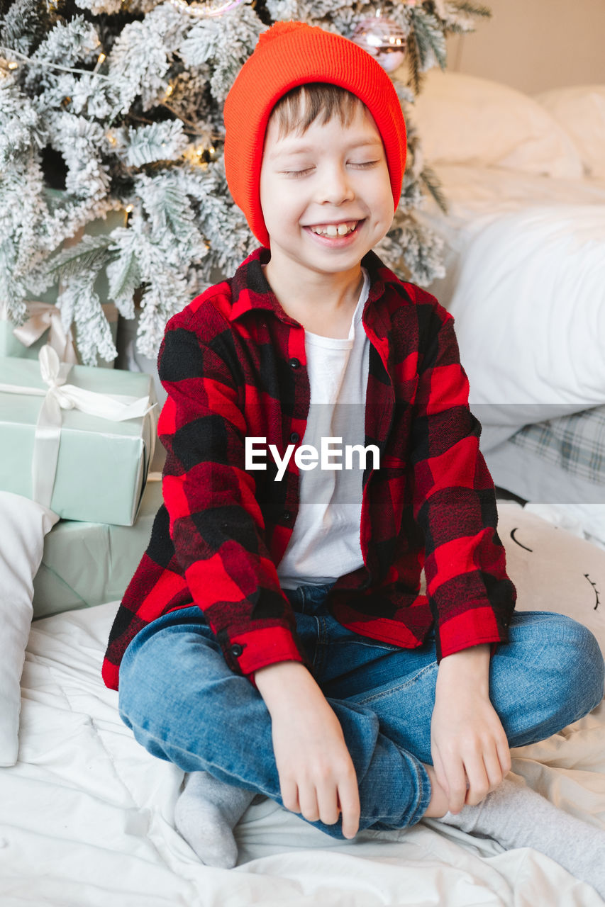 Boy in a red cap and a plaid shirt sits near a christmas tree with gifts on the floor and smiles