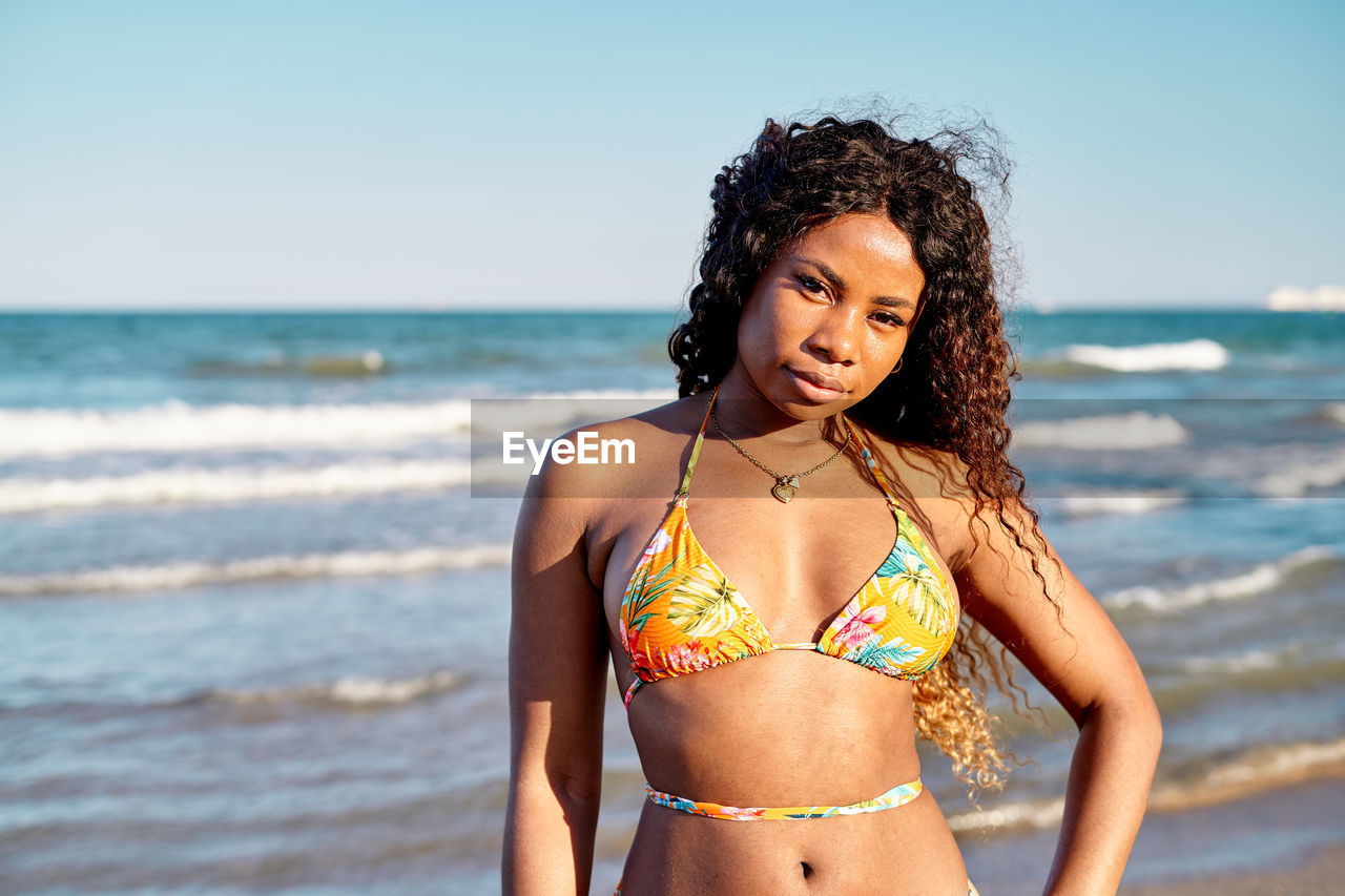 Happy african american female tourist in bikini holding hand on waist and looking at camera with smile while standing on beach near waving sea on summer day