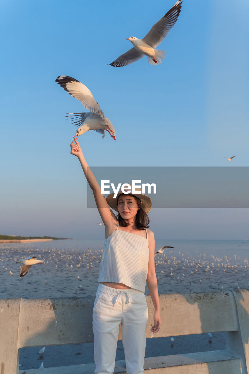 Beautiful woman wearing hat feeding seagull while standing against sky