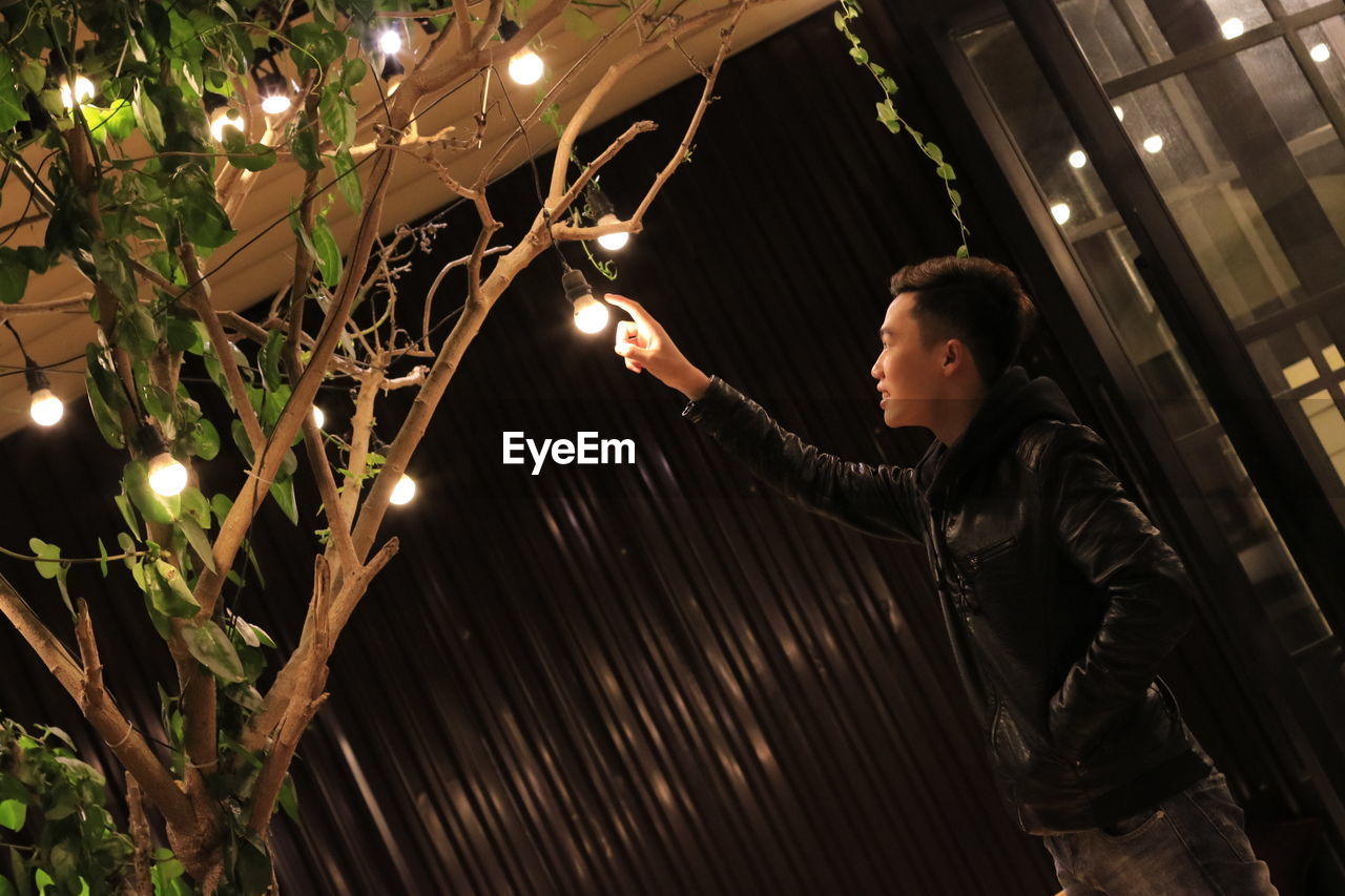 Young man touching electric bulb hanging from tree