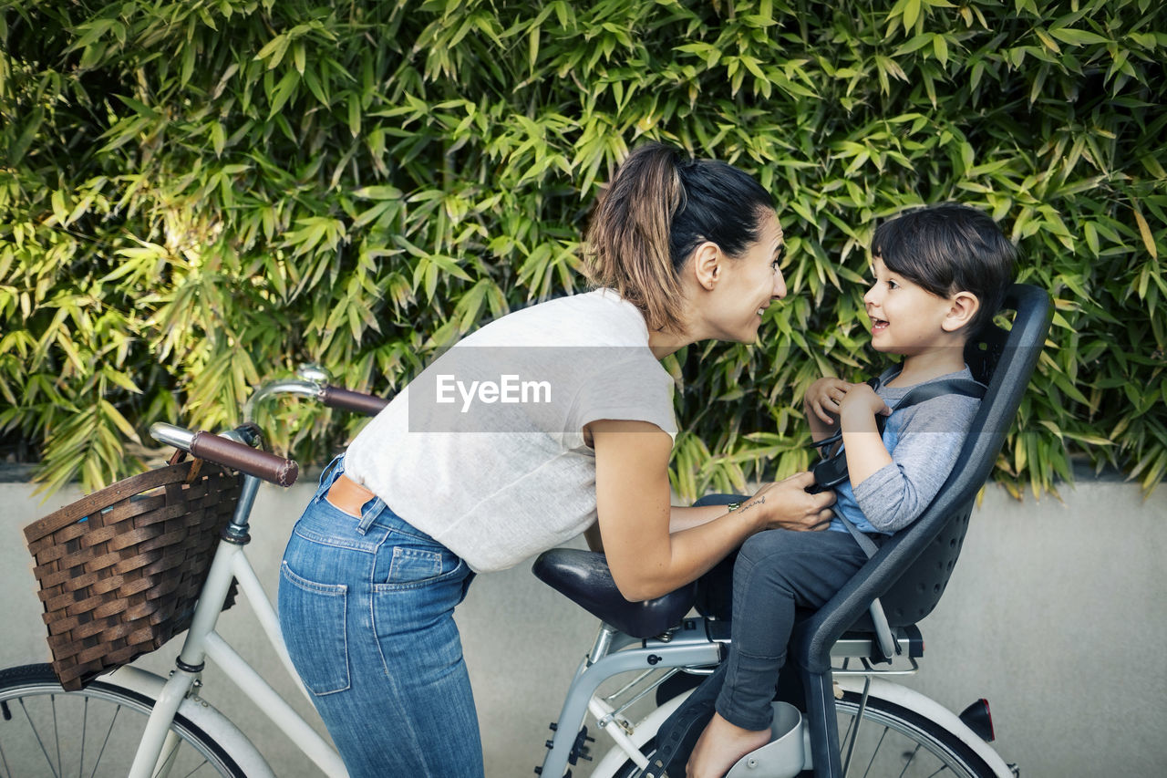 Happy woman looking at son sitting on back seat of bicycle against plants