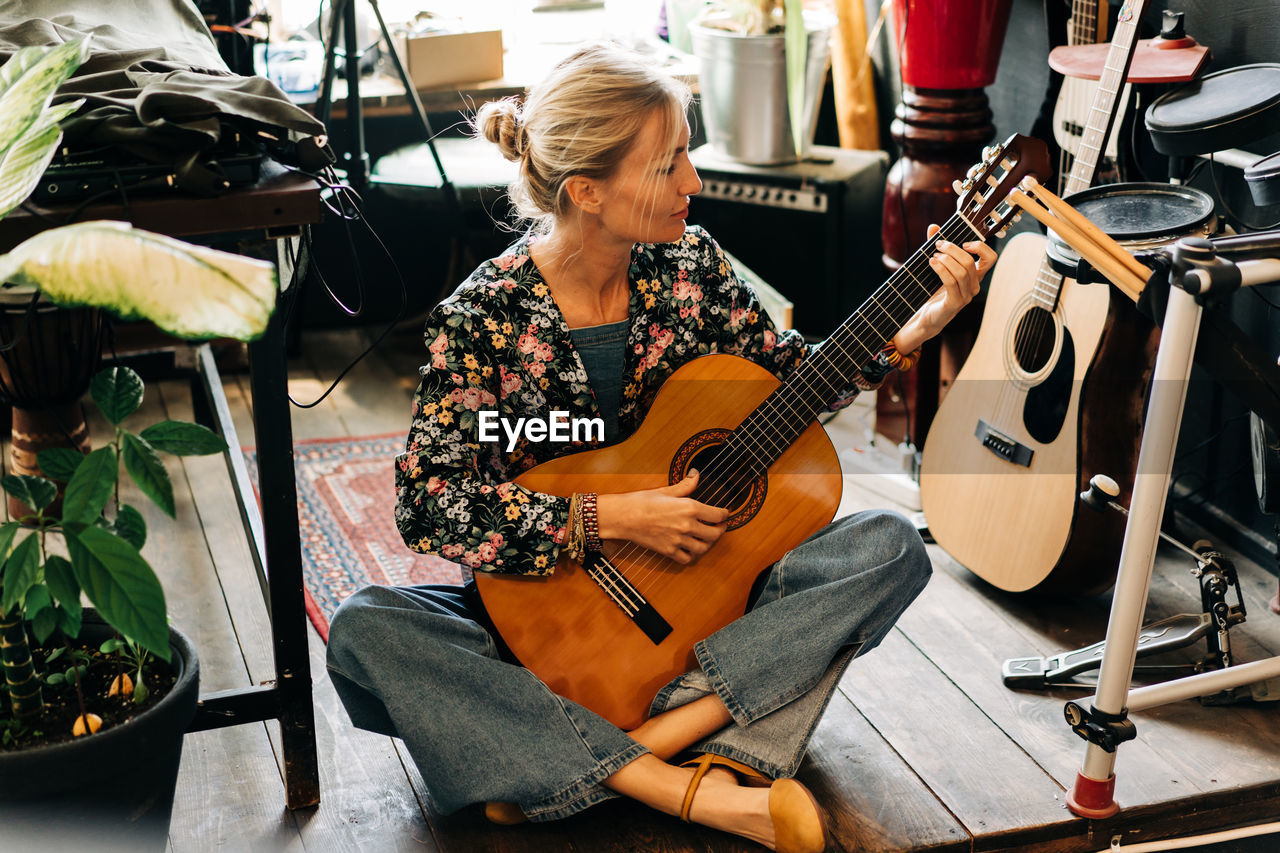 A woman plays an acoustic guitar while sitting on a stage among musical instruments.