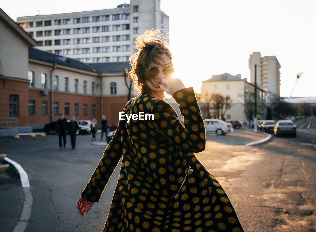 Portrait of young woman wearing sunglasses and overcoat on city street during sunset