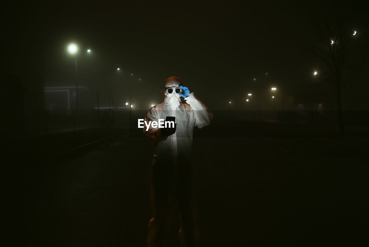 MAN STANDING ON STREET AGAINST ILLUMINATED LIGHT AT NIGHT