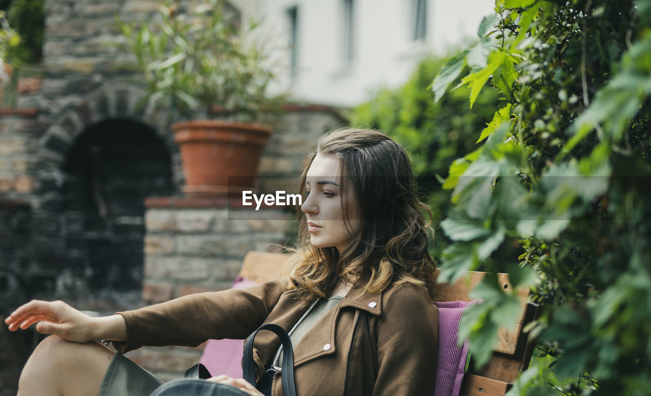 Thoughtful young woman sitting on bench by plant